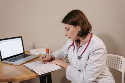 Médica escreve uma receita em um consultório, com um laptop sobre a mesa.