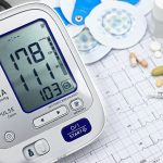 Monitor digital de pressão arterial, traçado de eletrocardiograma, comprimidos e instrumentos médicos sobre uma superfície clara.