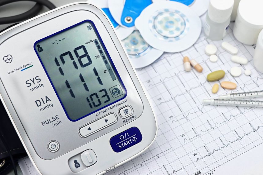 Monitor digital de pressão arterial, traçado de eletrocardiograma, comprimidos e instrumentos médicos sobre uma superfície clara.