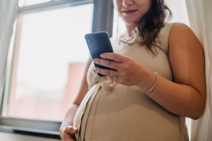 Gestante não identificada consulta notificações no celular próxima a uma janela.